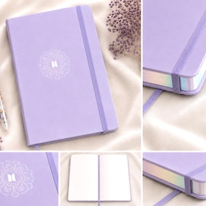 May include: A light purple notebook with a matching elastic closure and a white mandala-style logo on the cover. The notebook features iridescent page edges and comes with a coordinating purple and clear pen with internal graphic details.