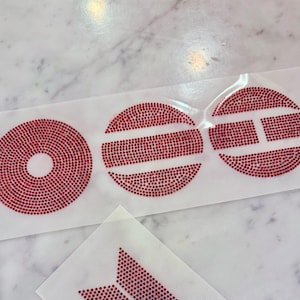 May include: A set of red rhinestone iron-on transfers on a clear adhesive backing. The designs include a solid circle, a circle with a horizontal bar, a circle with a vertical bar, and a partial triangular shape made of small red crystals.