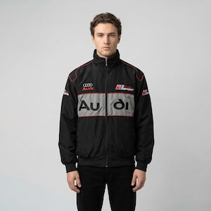 May include: Black zip-up jacket with red trim and the Audi logo. The jacket features a gray horizontal stripe across the chest with the word "Audi" in large letters. The Audi Sport logo is also visible.