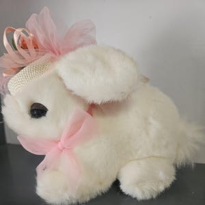 May include: A plush white bunny rabbit toy with a pink bow and a decorative hat. The bunny has black eyes and a fluffy tail. The hat is adorned with pink tulle and ribbons.