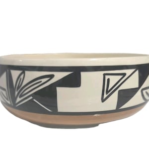 May include: A ceramic bowl with a cream-colored rim and a brown base. The bowl features a black and white geometric design with triangles and leaf-like shapes. The bowl's design is a striking contrast against the cream and brown colors.
