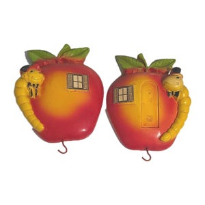 May include: Two apple-shaped wall decorations. Each apple is red and yellow with a green leaf and a small window. A yellow worm with a top hat and bow tie is attached to each apple. Each apple has a small hook.