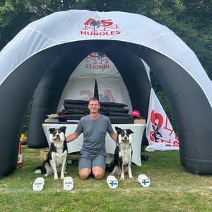 May include: A large white and black inflatable arch tent with the text AS HURDLES printed on the top. Two black and white border collie dogs sit on the grass, each holding a white dumbbell in their mouth, with two additional dumbbells on the ground.