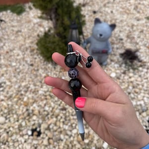 May include: A black and silver pen with decorative beads. The pen has a gradient design, transitioning from black to silver. The beads include a leopard print bead, and black spherical beads. A small charm with black cherry-shaped beads is attached.