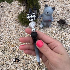 May include: A decorative pen with a black and white checkered heart-shaped bead, silver beads, and a black foam bead. The pen has a gradient design, transitioning from black to gray. The pen is held in a hand with pink nail polish.