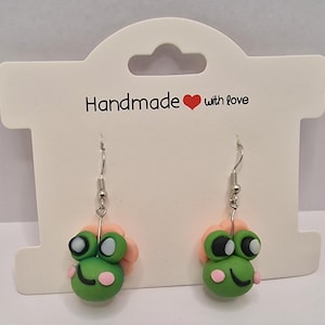 Op de afbeelding: Handgemaakte kikker oorbellen met groene lichamen, zwart-witte ogen en roze accenten. De oorbellen hangen aan zilverkleurige haken en worden gepresenteerd op een kaart met de tekst "Handmade with love."