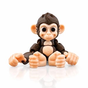 May include: A small articulated monkey figurine with dark brown fur texture on the head, arms, and back, contrasted by light beige skin on the face, ears, chest, hands, and feet. The figure features large black eyes and a seated posture.