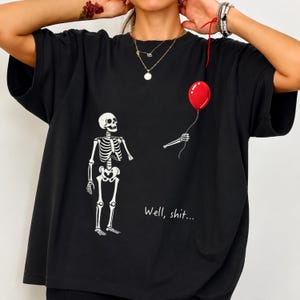 Peut inclure: Un t-shirt noir avec un motif blanc représentant un squelette debout à côté de son propre bras détaché, tenant un ballon rouge vif. Le texte "Well, shit..." est imprimé en blanc sous le squelette. Le t-shirt a des manches courtes.