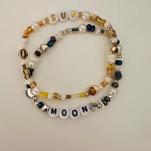 May include: Two beaded bracelets featuring white round beads with gold lettering spelling SUN and black lettering spelling MOON. The bracelets are composed of a mix of white faux pearls, metallic gold, silver, yellow, and dark blue glass beads.