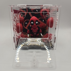 Puede incluir: Una vitrina de acrílico transparente con una figura de Deadpool. La figura es roja y negra, con una máscara y un disfraz detallados. La vitrina contiene detalles en rojo y un fondo temático de Deadpool. El panel frontal está abierto.