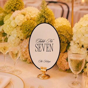 May include: An oval table number sign with a black border, white background, and black text reading Table No. SEVEN and The Kelseys. The sign is held upright by a small, circular gold metal stand, positioned on a table with floral arrangements.