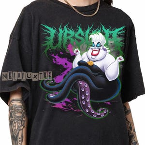 May include: Black t-shirt featuring a cartoon illustration of Ursula from The Little Mermaid. The design includes the word "URSULA" in green and purple, with the character's image in the center. The shirt also has the text "NELLOLUXTEE".