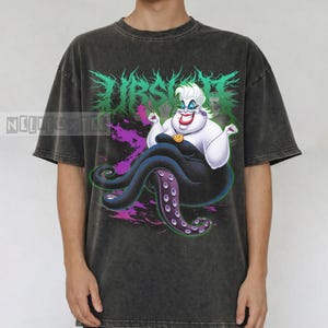 May include: Dark gray t-shirt featuring a graphic of Ursula from The Little Mermaid. The design includes the word "Ursula" in green, a purple and pink background, and Ursula's likeness. The shirt is a short-sleeved, casual style.