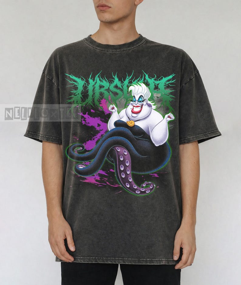 May include: Dark gray t-shirt featuring a graphic of Ursula from The Little Mermaid. The design includes the word "Ursula" in green, a purple and pink background, and Ursula's likeness. The shirt is a short-sleeved, casual style.
