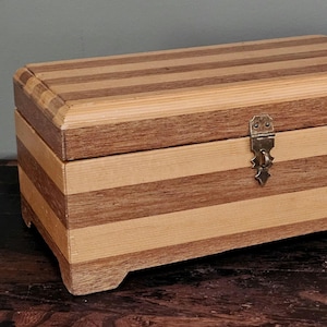 May include: A rectangular wooden box featuring alternating horizontal stripes of light and dark wood grain. The lid is hinged and secured with a central brass-coloured metal latch. The box is supported by four small, integrated wooden feet.