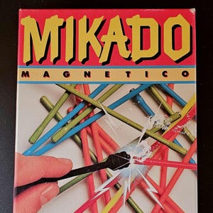 Peut inclure: Une boîte de jeu rectangulaire pour Mikado Magnetico avec le titre en lettres jaunes sur fond rouge. La couverture montre un tas de bâtonnets bleus, verts, jaunes et rouges avec un outil magnétique noir attirant un bâtonnet rouge avec un graphique d'étincelle blanche. Un logo éléphant Jumbo rose est en bas à droite.