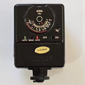 Peut inclure: Un flash électronique noir pour appareil photo doté d'un cadran circulaire avec réglages ASA, échelles d'ouverture et marquages de distance. L'appareil comprend un interrupteur coulissant pour les modes auto, manuel et arrêt, et un autocollant jaune PASSED.