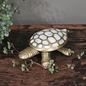 May include: A decorative turtle figurine crafted from brass with a polished finish. The shell features an inlaid pattern of smooth, white, iridescent mother-of-pearl segments set within the gold-toned metal framework of the turtle's back.