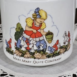 Peut inclure: Un mug en céramique blanche orné d'une illustration colorée représentant une personne aux cheveux blonds vêtue d'une tenue rouge et bleue, arrosant des fleurs avec un arrosoir vert. Le texte "MARY, MARY, QUITE CONTRARY" est imprimé en noir.