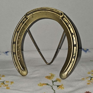 May include: A gold-toned metal horseshoe stand featuring the embossed text TRIPLE CROWN on the left side and the number 3 on the right side. The horseshoe is supported by a thin, triangular metal wire frame attached to the back of the object.