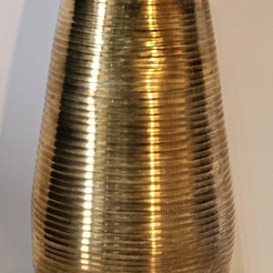 Peut inclure: Un vase doré avec un motif texturé et nervuré. Le vase a une base large qui se rétrécit vers le haut jusqu'à une ouverture plus étroite. La finition métallique reflète la lumière, créant des reflets et des ombres. C'est un objet décoratif.