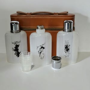May include: A vintage drinks set with three white glass flasks, silver caps, and dog illustrations. The flasks have text: "One Scotch after another", "Hm bourbon!", and "Comin thru the rye". The set includes a small metal cup and a stack of clear plastic cups. A brown leather case is in the background.