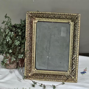 May include: An antique gold-coloured picture frame with a rectangular shape. The frame features detailed, ornate patterns around the edges. The frame is empty, with a dark, reflective surface. It is displayed on a white tablecloth, with greenery in the background.