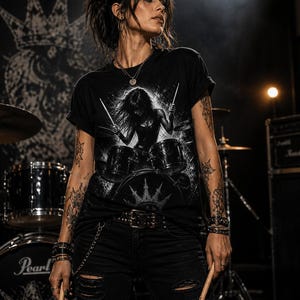 May include: Black t-shirt with a drummer graphic. The design shows a drummer playing a drum kit with a crown above the drums. The person in the image is wearing black ripped jeans, a belt, and bracelets, holding drumsticks.
