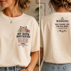 Op de afbeelding: Een crèmekleurig t-shirt met een afbeelding op de voorkant van gestapelde boeken, een bril en rozen met de tekst "MY BOOK BOYFRIEND PUTS THE CAP BACK ON THE TOOTHPASTE". De achterkant toont een waarschuwingssymbool en de tekst "WARNING MAY IGNORE YOU FOR ONE MORE CHAPTER".