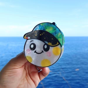Puede incluir: Un accesorio para el cabello circular que presenta una cara de dibujos animados con grandes ojos negros y una boca sonriente. La parte superior es una gorra negra con estrellas doradas, mientras que la parte inferior es blanca con patrones circulares amarillos y rosas.
