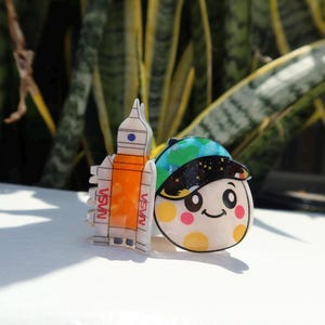 May include: A white and orange rocket ship hair claw clip with the text NASA printed in red, positioned next to a round hair clip featuring a smiling face character wearing a blue, green, and black space-themed cap with yellow and pink polka dots.