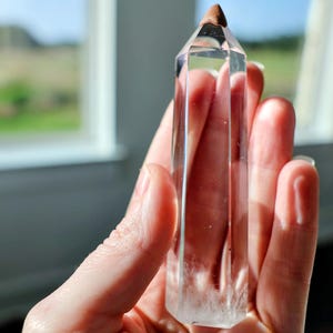 May include: A clear, hexagonal quartz crystal point with a smooth, polished surface and a distinct, pointed tip. The crystal is transparent, showing internal natural inclusions and a solid, faceted structure that tapers to a sharp apex.