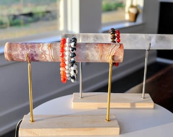 Könnte beinhalten: Zwei Schmuckständer mit hellen Holzsockeln und Metallstangen. Der vordere Ständer hat eine zylindrische Stange mit violetten, weißen und orangefarbenen gebänderten Steinmustern, an der drei Perlenarmbänder in Orange, Weiß sowie Schwarz-Weiß hängen.