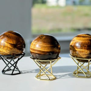 Puede incluir: Tres esferas de piedra ojo de tigre pulidas con características bandas chatoyantes de color marrón dorado. Cada esfera descansa sobre un soporte de metal geométrico, con un soporte negro a la izquierda y dos soportes dorados en el centro y a la derecha.