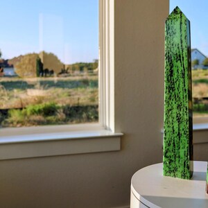 May include: Two tall, pointed stone obelisks stand upright. The taller obelisk is bright green with dark black streaks. The shorter obelisk features a mix of vibrant green and deep reddish-brown patches with dark black inclusions throughout the stone.