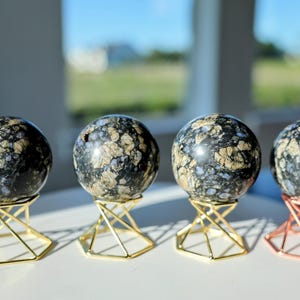 Peut inclure: Quatre pierres sphériques polies présentant une base noire foncée avec des inclusions tachetées de couleur beige et grise. Chaque sphère repose sur un support métallique géométrique, trois dorés et un cuivré soutenant les pierres.