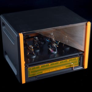 May include: Black and orange display case with a clear front panel. The case contains several miniature figurines on a black shelf. The text "SQUAD GAMES PROTECTS" is visible on the front of the case.