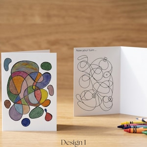 Peut inclure: Deux cartes de vœux pliées blanches. La carte de gauche présente un motif abstrait coloré avec des lignes courbes entrelacées et des formes remplies de bleu, vert, jaune et rouge. La carte de droite montre un dessin au trait noir avec le texte Now your turn.