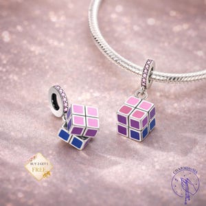 May include: Two silver charm pendants featuring miniature Rubik's Cubes in shades of pink, purple, and blue. One charm is attached to a silver bracelet. The charms have a geometric design and are accented with pink gemstones.