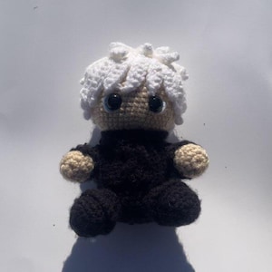 Könnte beinhalten: Handgemachte Häkelpuppe mit weißer, strukturierter Frisur und großen blauen Augen. Die Puppe trägt ein schwarzes Outfit mit beigefarbenen Armen und Beinen. Die Puppe sitzt.