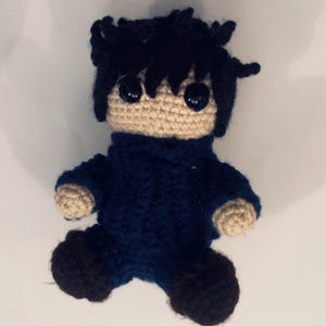 May include: A handmade crochet doll with black hair and large black eyes. The doll is wearing a dark blue outfit with tan hands and face. The doll is sitting with its legs bent. The doll is a handmade item.