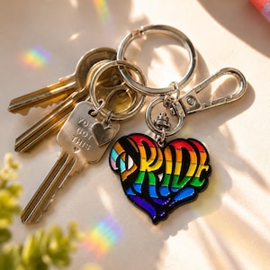 May include: A silver-toned metal keychain featuring a heart-shaped charm with rainbow-colored enamel and the word PRIDE in bold black lettering. Attached is a smaller silver heart charm engraved with the text YOU GOT THIS and several keys.