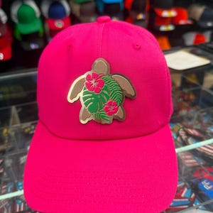 May include: A bright pink baseball cap featuring a centered patch of a brown sea turtle. The turtle shell is decorated with green monstera leaves and two vibrant pink hibiscus flowers. The cap has a structured crown and a curved brim.