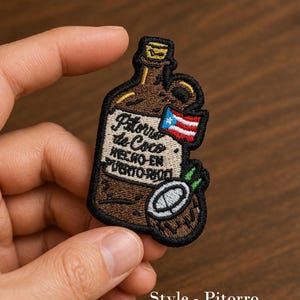 May include: Embroidered patch in the shape of a brown bottle with the text "Pitorro de Coco HECHO EN PUERTO RICO". Features a Puerto Rican flag and a coconut design. The patch is outlined in black.