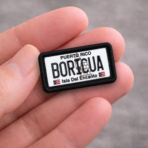 May include: A rectangular patch with a black border, featuring the text "PUERTO RICO" above "BORICUA" in bold white letters. Below, it reads "Isla Del Encanto" with a small Puerto Rican flag on each side.