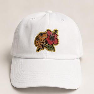 May include: White baseball cap featuring an embroidered design of a frog and a red hibiscus flower. The frog is brown with gold accents, and the flower has a green leaf. The cap has a curved brim and an adjustable strap.