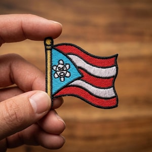 May include: Embroidered patch of the Puerto Rican flag, featuring red and white stripes with a blue triangle and white star. The patch is held between fingers against a wooden background. The flag is a symbol of Puerto Rican pride.