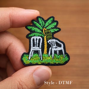 May include: Embroidered patch featuring a palm tree with two white chairs beneath it. The palm tree has green fronds and a yellow trunk with a bunch of bananas. The patch has a black border and the text "Style - DTMF".