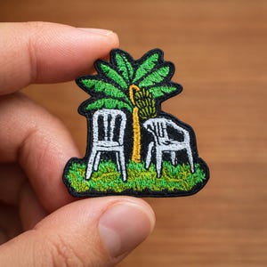 May include: Embroidered patch featuring a green palm tree with a yellow trunk and a cluster of bananas. Two white chairs are positioned beneath the tree on a green grassy area. The patch has a black border.
