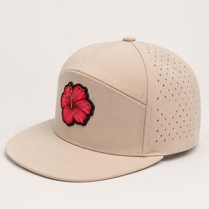 May include: A beige baseball cap with a flat brim and a perforated side panel. The front panel features a prominent embroidered patch of a vibrant red hibiscus flower with black outlines and a dark border, centered on the smooth beige fabric.